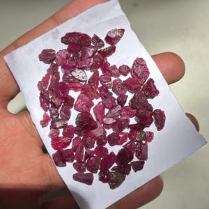 Peut inclure: Collection de pierres précieuses de rubis bruts. Les rubis non taillés varient en taille et en forme, affichant une teinte rouge profond avec des zones roses plus claires et cramoisies plus foncées. Les pierres sont présentées sur une surface blanche.