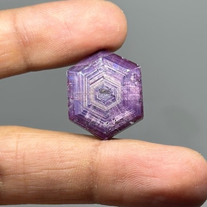Puede incluir: Cristal hexagonal púrpura facetado, sostenido entre los dedos. La superficie del cristal muestra un complejo patrón geométrico con un diseño central. El color es un violeta intenso con tonos más claros dentro de las facetas, creando un efecto brillante.