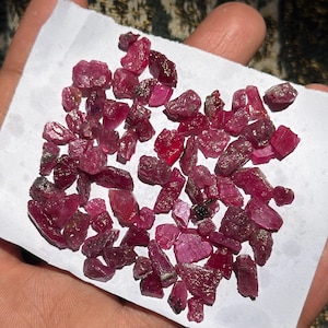 Può includere: Una collezione di piccole pietre preziose di rubino grezze e non lucidate in varie tonalità di rosa intenso e rosso. Questi frammenti minerali naturali e irregolari presentano superfici sfaccettate e texture cristalline.
