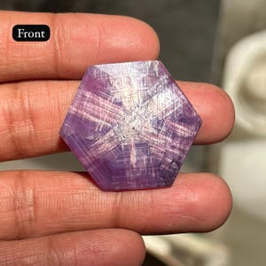 May include: A hexagonal, purple gemstone with a complex, linear pattern radiating from the center. The word "Front" is visible in the upper left corner. The gemstone is held between two fingers.