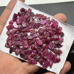Könnte beinhalten: Eine Sammlung roher, ungeschliffener Rubin-Edelsteine. Die Rubine sind tiefrot und weisen Farb- und Texturvariationen auf. Sie werden auf einer weißen Oberfläche präsentiert, die ihre natürlichen, unregelmäßigen Formen hervorhebt.