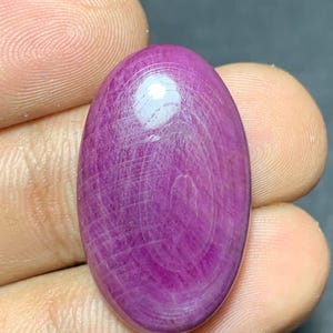 May include: An oval-shaped cabochon gemstone with a deep magenta and purple hue. The surface displays a distinct, natural cross-hatched pattern of fine lines and internal inclusions, with a smooth, polished finish that reflects light.