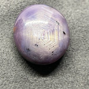 May include: A round, cabochon-cut star sapphire gemstone with a translucent lavender and light purple hue. The surface displays a distinct, centered hexagonal star pattern with light-reflecting rays and several small, dark, natural mineral inclusions.