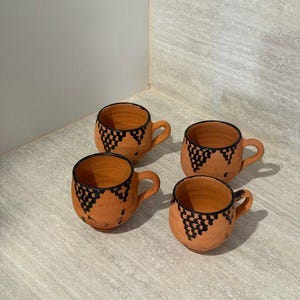 Peut inclure: Quatre tasses en terre cuite avec de petites anses. Chaque tasse présente un motif géométrique noir sur le bord. Les tasses sont de couleur orange chaud et semblent faites à la main.