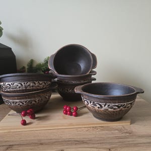 Könnte beinhalten: Eine Sammlung handgefertigter, dunkelbrauner Keramikschalen mit Griffen. Jede Schale ist mit einem weißen Blumenmuster am Rand verziert. Die Schalen sind auf einem Holzbrett angeordnet, mit roten Beeren.