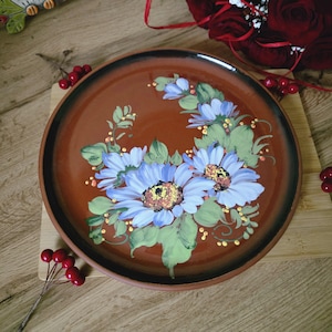 Może przedstawiać: Okrągły, ręcznie malowany ceramiczny talerz z brązową podstawą i czarną obwódką. Talerz zdobi kwiatowy wzór z niebieskich kwiatów z żółtymi środkami i zielonymi liśćmi. Talerz jest prezentowany na drewnianej powierzchni.