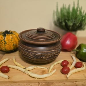 Può includere: Un vaso di ceramica marrone con coperchio, con un design strutturato. Il vaso è circondato da una piccola zucca gialla, peperoni rossi e verdi, fagiolini e pomodorini su un tagliere di legno. Una pianta verde è sullo sfondo.