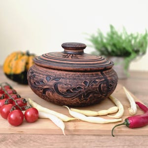 Può includere: Un vaso di ceramica marrone con coperchio, con disegni floreali intagliati. Il vaso è circondato da verdure fresche, tra cui pomodorini rossi, fagiolini gialli e un peperoncino rosso. Una piccola zucca è sullo sfondo.