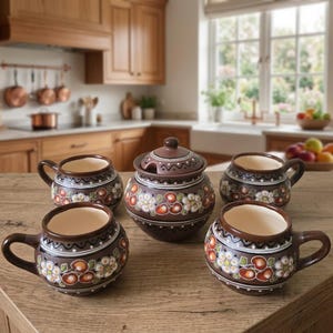 Könnte beinhalten: Ein Set aus vier braunen Keramiktassen und einer passenden Zuckerdose mit Deckel, jeweils mit handgemaltem Blumenmuster aus weißen Blütenblättern, grünen Blättern und rotbraunen Akzenten, verziert mit einem dunkelbraunen Rand.