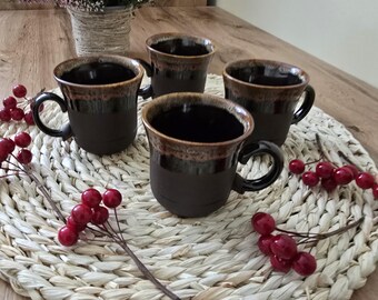 Può includere: Quattro tazze in ceramica marrone scuro con bordo e manico marrone a contrasto sono disposte su un sottobicchiere intrecciato. Le tazze sono vuote e hanno una finitura lucida. Nell'immagine sono presenti anche bacche rosse decorative e un vaso di fiori viola.