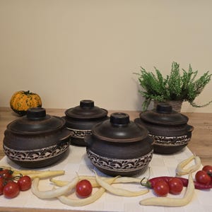 Może przedstawiać: Cztery ciemnobrązowe ceramiczne garnki do gotowania z pokrywkami, ułożone na białej tkaninie. Każdy garnek ma kremowy ozdobny pasek. Warzywa, w tym pomidory, fasola i papryka, są ułożone przed garnkami. Mała dynia i roślina doniczkowa znajdują się w tle.