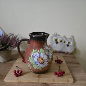 Könnte beinhalten: Ein brauner Keramikkrug mit Henkel, verziert mit handgemalten Blumenmustern in Blau, Weiß und Gelb. Der Krug steht auf einem Holzschneidebrett, mit roten Beeren verstreut. Im Hintergrund befindet sich eine dekorative Eule.