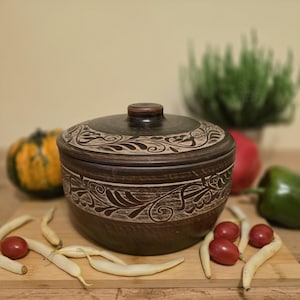 Può includere: Una ciotola marrone in ceramica con coperchio e motivo floreale. La ciotola è circondata da verdure, tra cui pomodori, peperoni verdi e una piccola zucca. La ciotola è su un tagliere di legno.