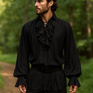 May include: Black long-sleeve shirt with a ruffled front and cuffs. The shirt has a lace-up neckline and is paired with a black belt. The image is set against a blurred green background.