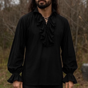 May include: Black long-sleeve shirt with a ruffled front and cuffs. The shirt has a collared neckline and a lace-up detail at the chest. The sleeves are long and loose, with ruffled cuffs at the wrists. The shirt is made of a soft, flowing fabric.