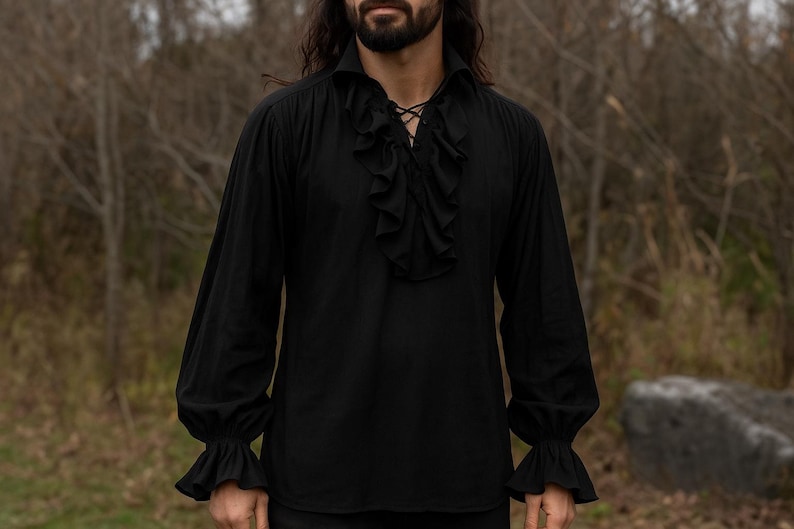 May include: Black long-sleeve shirt with a ruffled front and cuffs. The shirt has a collared neckline and a lace-up detail at the chest. The sleeves are long and loose, with ruffled cuffs at the wrists. The shirt is made of a soft, flowing fabric.