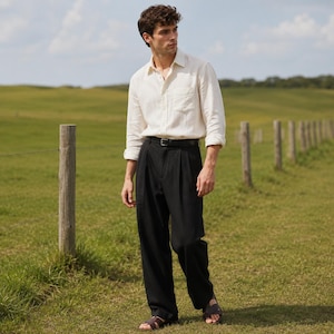 May include: A long-sleeved, off-white button-down shirt with a chest pocket, paired with loose-fitting black trousers featuring a pleated front and a black belt. The outfit is completed with dark brown leather sandals worn on the feet.