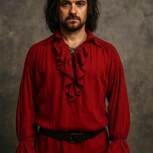 May include: A red long-sleeved shirt with a ruffled front and a black belt. The shirt has a loose fit with flared sleeves. The person in the image has long, dark hair and a beard.