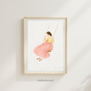 Puede incluir: Una impresión artística enmarcada que presenta una ilustración en acuarela de una persona con vestimenta tradicional coreana en un columpio. La persona lleva una parte superior amarilla y una falda rosa fluida. La impresión tiene un marco de madera clara y el texto "studiodearsoul" en la parte inferior.