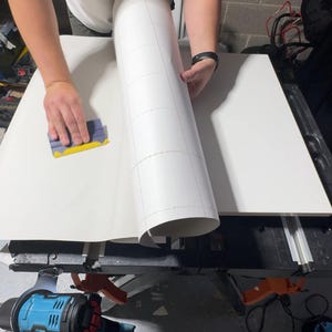 May include: A person is applying a white vinyl sheet with a grid pattern to a white surface. A yellow and blue squeegee is used to smooth the vinyl. Tools and equipment are visible.