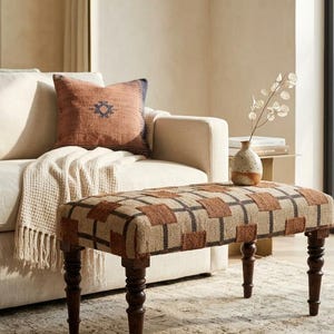 May include: A rectangular bench with a brown and tan checkered pattern fabric seat. The bench has dark wooden legs with a turned design. A beige sofa, throw pillow, and vase with a plant are in the background.