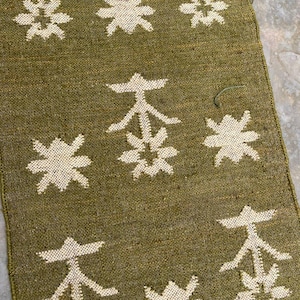 May include: A rectangular olive green textile runner featuring a repeating pattern of cream-colored woven motifs. The design consists of alternating stylized floral shapes and geometric symbols arranged in a grid across the fabric surface.
