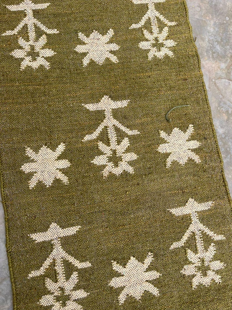 May include: A rectangular olive green textile runner featuring a repeating pattern of cream-colored woven motifs. The design consists of alternating stylized floral shapes and geometric symbols arranged in a grid across the fabric surface.