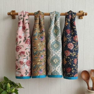 May include: Four patterned kitchen towels hang from a wooden rail. The towels display floral designs in pink, brown, teal, and navy blue. Each towel has a contrasting blue border. The towels are a decorative addition to any kitchen, with a range of floral patterns.
