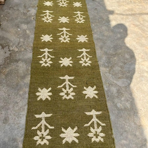 May include: A long, rectangular olive green runner rug featuring a repeating pattern of white star-like geometric shapes and stylized tree motifs. The rug has a woven texture and is finished with short, dark gray fringe at both ends.