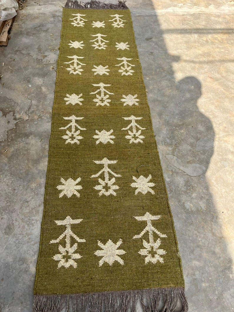 May include: A long, rectangular olive green runner rug featuring a repeating pattern of white star-like geometric shapes and stylized tree motifs. The rug has a woven texture and is finished with short, dark gray fringe at both ends.