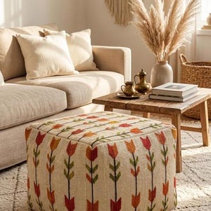 May include: A square ottoman with a beige fabric featuring a repeating pattern of red, orange, and green tulip-like designs. The ottoman is in a living room setting with a beige sofa, wooden table, and decorative items.