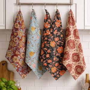 May include: Four decorative kitchen towels with floral patterns hang from a metal bar. The towels feature designs in burgundy, light blue, black, and beige, each with a unique floral print. The towels are hanging in front of a white tiled wall.