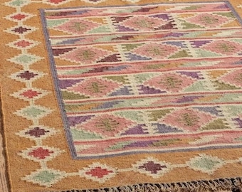May include: A rectangular area rug with a geometric pattern. The rug features a border of diamond shapes in various colors, including red, green, and purple, set against a tan background. The central design includes horizontal stripes and diamond shapes in pink, purple, and green.