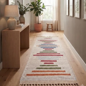 May include: A long, rectangular runner rug featuring a cream-colored base with a central pattern of horizontal stripes in shades of red, green, blue, and orange. The rug is finished with decorative fringe tassels along both of its short ends.