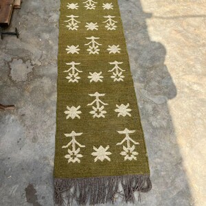May include: Olive green runner rug with a repeating white floral pattern. The rug has a fringed edge and is made of a woven material. The rug is rectangular and appears to be handmade.