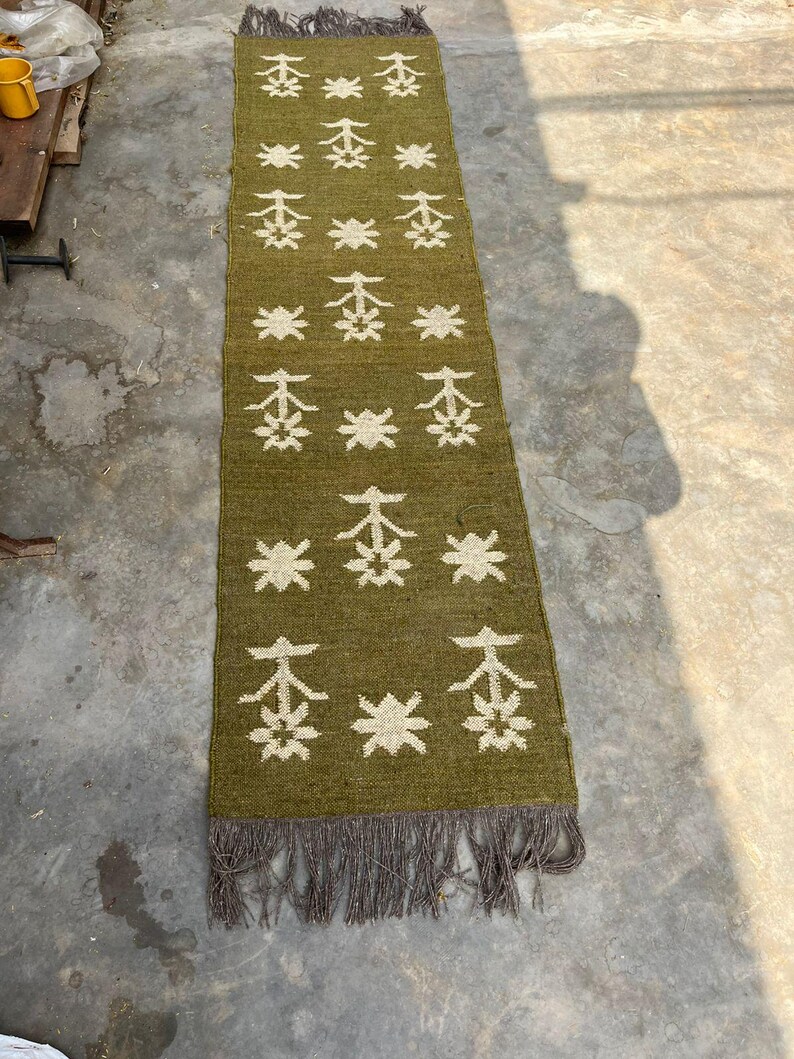 May include: Olive green runner rug with a repeating white floral pattern. The rug has a fringed edge and is made of a woven material. The rug is rectangular and appears to be handmade.
