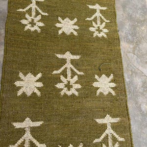 May include: A rectangular woven runner in olive green featuring a repeating pattern of off-white geometric star-like shapes and stylized tree motifs arranged in a grid across the textured fabric surface.
