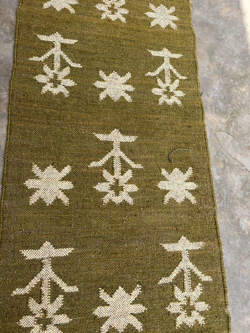 May include: A rectangular woven runner in olive green featuring a repeating pattern of off-white geometric star-like shapes and stylized tree motifs arranged in a grid across the textured fabric surface.