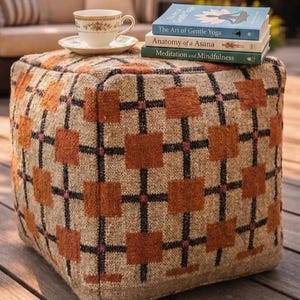 Puede incluir: Un otomán cuadrado con un patrón geométrico beige y naranja. Una pila de tres libros titulados "The Art of Gentle Yoga", "Anatomy of a Asana" y "Meditation and Mindfulness" se encuentra encima, junto con una taza y un platillo.