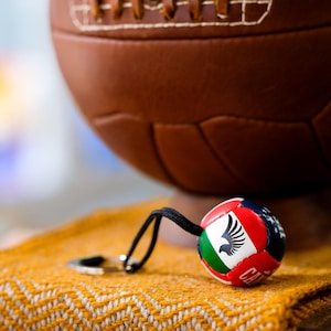May include: A brown leather vintage-style soccer ball with visible laces sits behind a small, round keychain. The keychain features a multi-colored design with a black eagle emblem, red, white, and green panels, and a black cord with a metal ring.