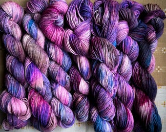 May include: A selection of hand-dyed yarn skeins in shades of purple, pink, and blue. The yarn is arranged in a row, displaying the colour variations. The skeins are on a light-coloured fabric with a floral pattern.