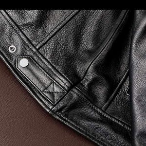 May include: Close-up of a black leather jacket, showcasing the textured grain and stitching details. The jacket features a snap closure and a quilted design element. The background is a brown leather surface.