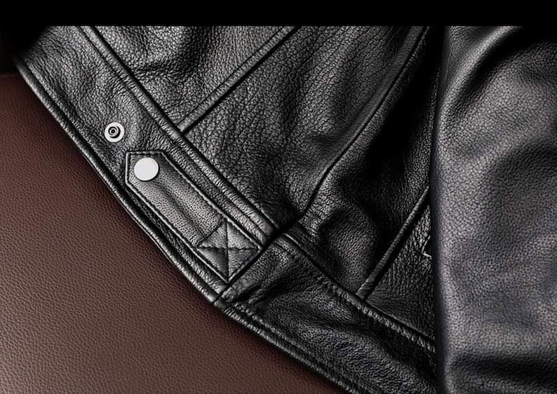 May include: Close-up of a black leather jacket, showcasing the textured grain and stitching details. The jacket features a snap closure and a quilted design element. The background is a brown leather surface.