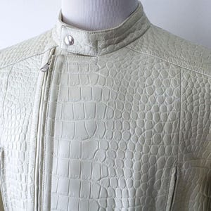May include: Cream-colored leather jacket with a textured, crocodile-like pattern. The jacket features a stand-up collar, a silver zipper, and a tailored fit. The design includes stitched details and a small pocket.