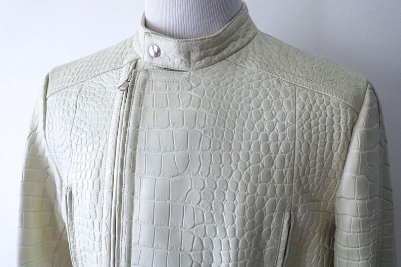 May include: Cream-colored leather jacket with a textured, crocodile-like pattern. The jacket features a stand-up collar, a silver zipper, and a tailored fit. The design includes stitched details and a small pocket.