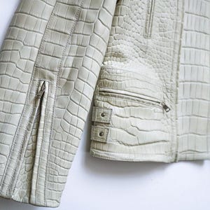 May include: Close-up of a light green leather jacket with a crocodile skin texture. The jacket features a zipper on the sleeve and a zippered pocket. The jacket has a silver lining.