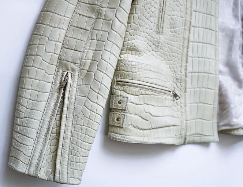 May include: Close-up of a light green leather jacket with a crocodile skin texture. The jacket features a zipper on the sleeve and a zippered pocket. The jacket has a silver lining.