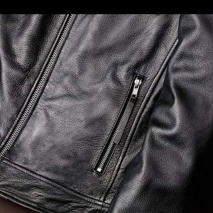 May include: Close-up of a black leather jacket with a textured surface. The jacket features a silver zipper closure and a zippered pocket on the chest. The leather appears to be supple and well-crafted.