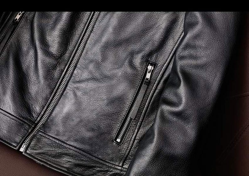 May include: Close-up of a black leather jacket with a textured surface. The jacket features a silver zipper closure and a zippered pocket on the chest. The leather appears to be supple and well-crafted.
