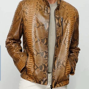 Może przedstawiać: Brązowa i beżowa kurtka ze wzorem skóry węża z zapięciem na zamek i dwiema przednimi kieszeniami. Kurtka ma stójkę i długie rękawy. Model ma na sobie szarą koszulę i białe spodnie.